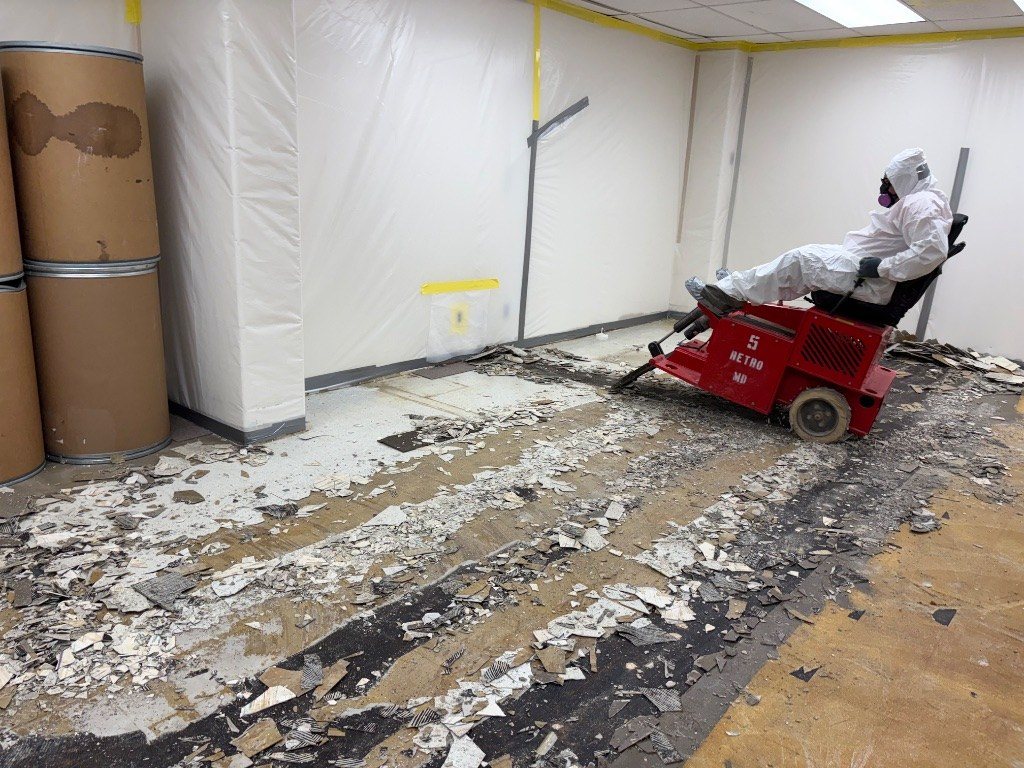 RETRO crew member in a protective suit and respirator removing floor tile with a RETRO ride-on scraper inside poly containment