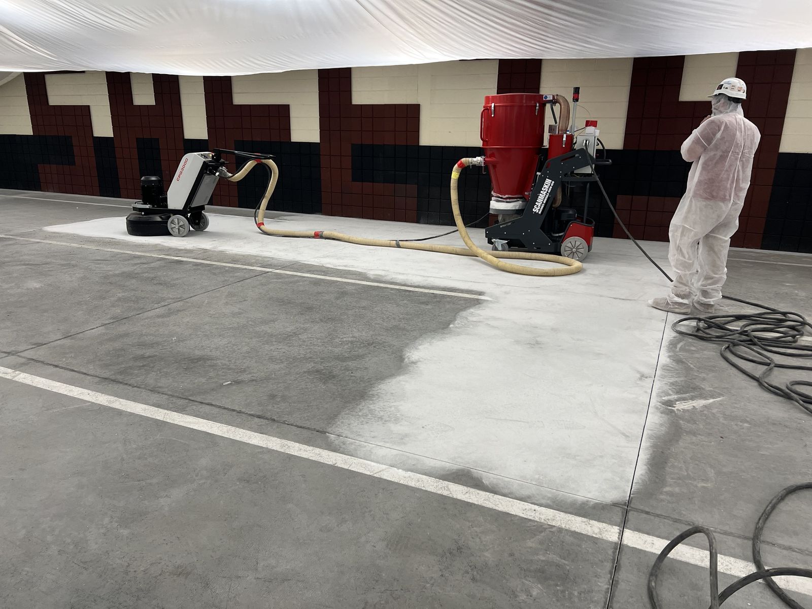 Ground section of a garage slab beside unground concrete, with a floor grinder, dust collector, and a RETRO crew member in a protective suit