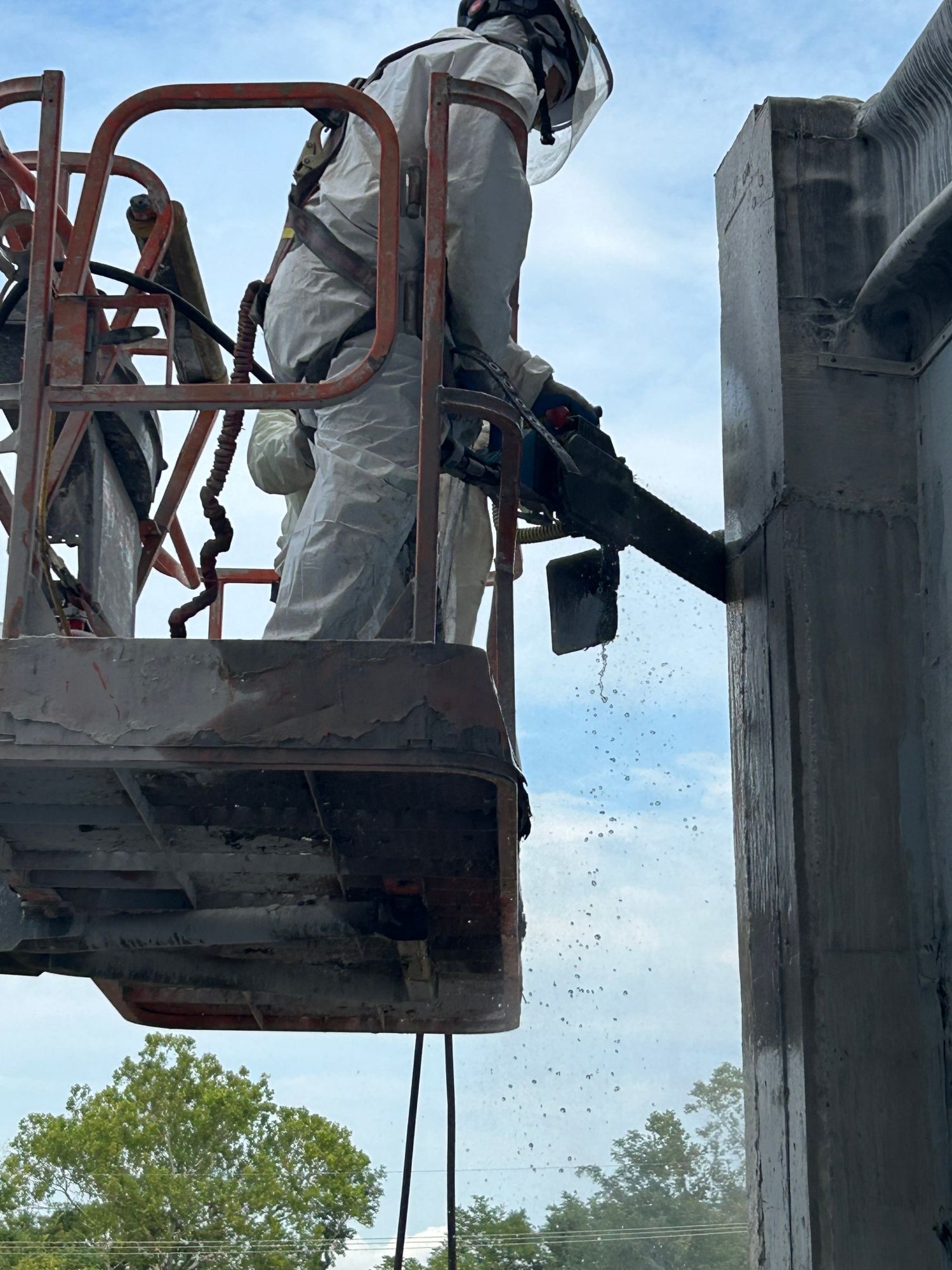 RETRO crew member in a protective suit and harness cutting a concrete wall with a diamond chain saw from a boom lift, water spraying from the cut