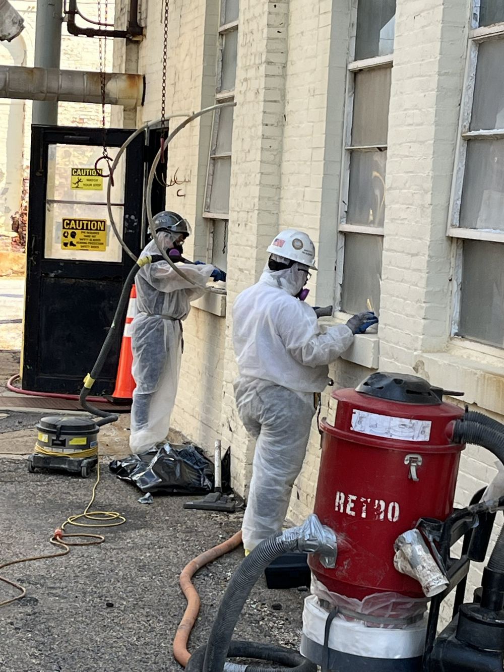 Two RETRO crew members in protective suits and respirators removing lead-based paint from window sills on a brick building, with a RETRO-labeled vacuum in front